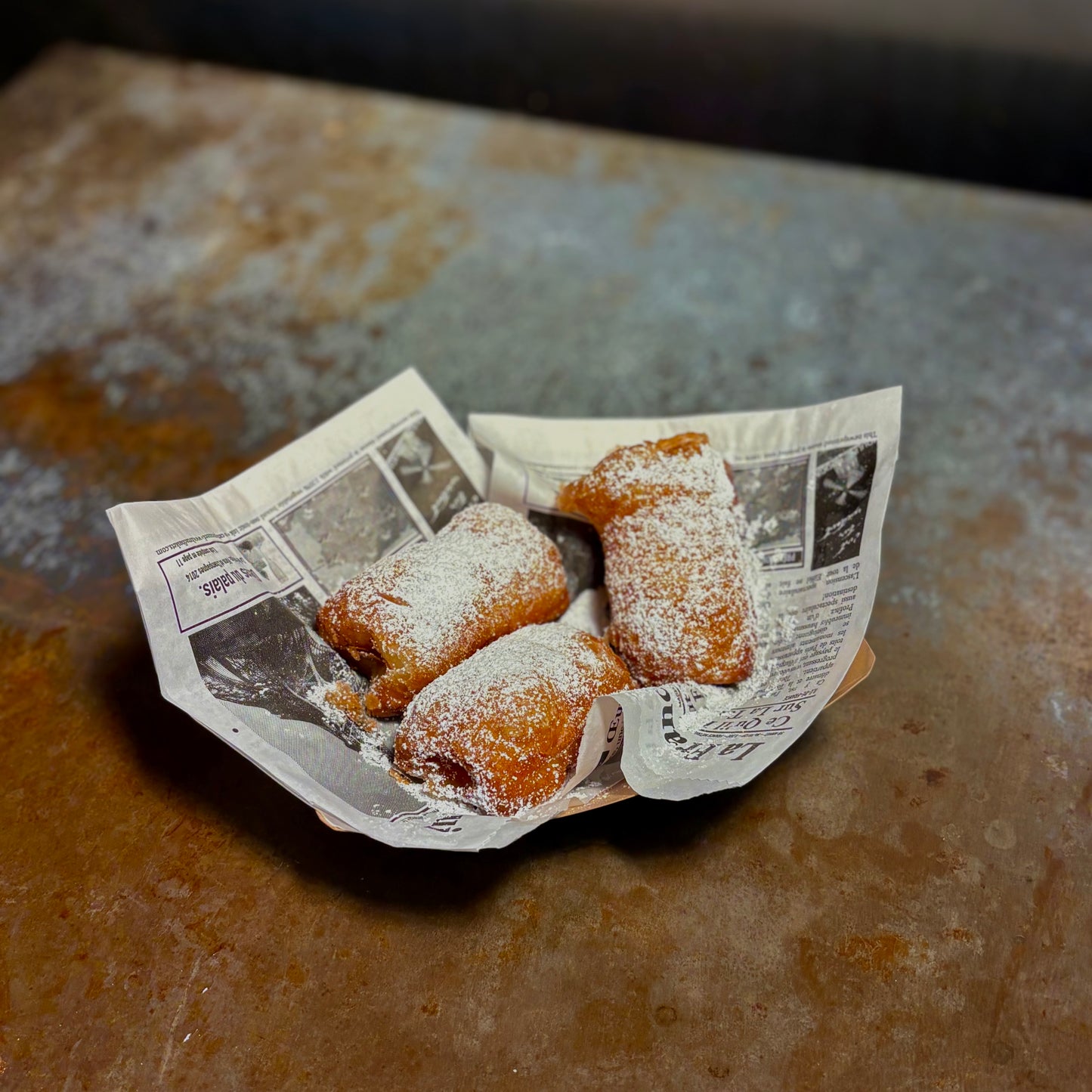 Three powdered sugar-covered pastries in a paper container on a rustic metal surface.