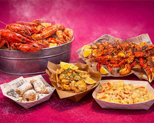A vibrant seafood spread from Boil Baby Boil featuring a large metal bucket of boiled crawfish, a basket of powdered beignets, a hot buttered lobster roll, and creamy lobster mac and cheese against a bright pink background.