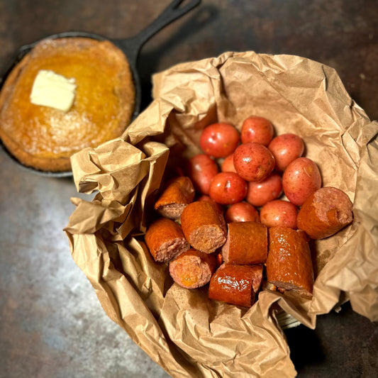 Spicy andouille sausage and red potatoes served in brown butcher paper, with honey butter cornbread in a cast iron skillet in the background.