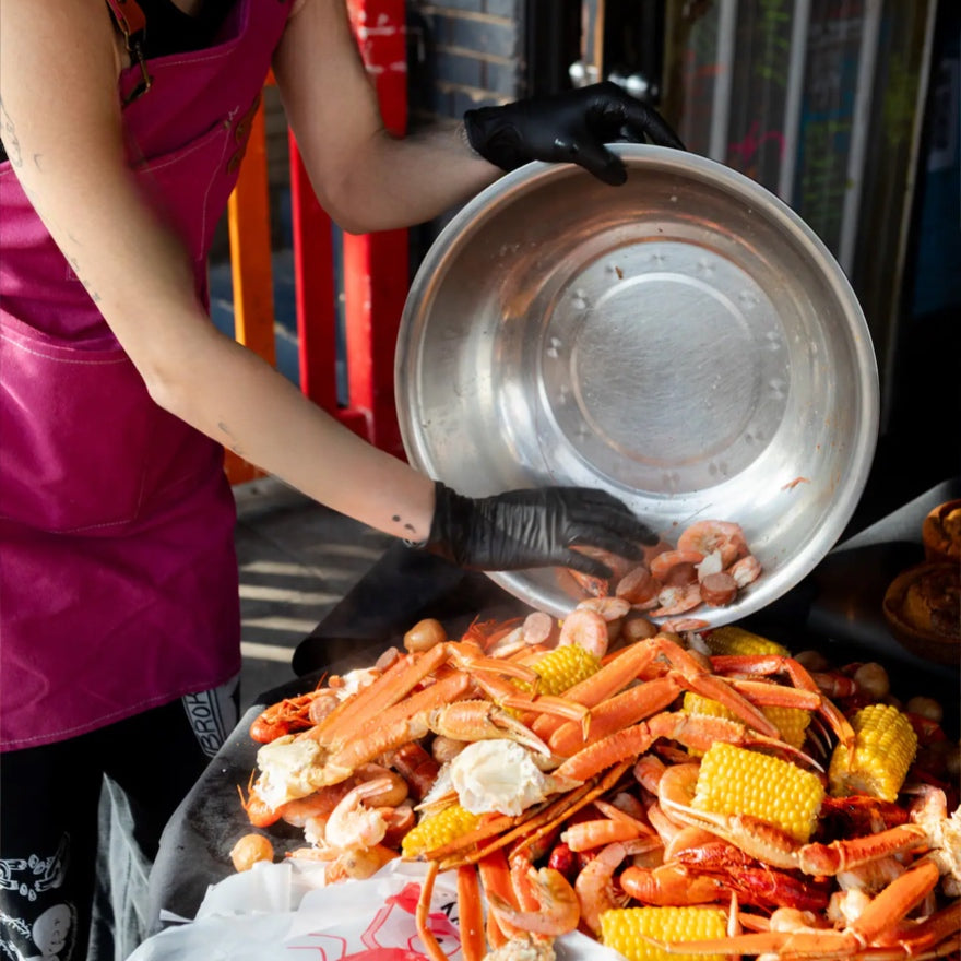 Person preparing seafood dish with shrimp and corn on a metal pan.