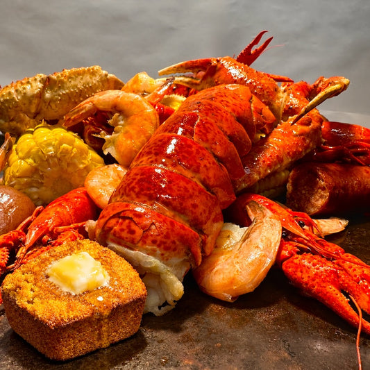A close-up of a cooked seafood boil or lobster meal on a dark skillet, featuring a large lobster tail, shrimp, crab, corn, and a bread cube topped with butter.