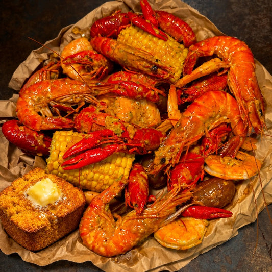 A close-up overhead shot of a seafood boil or crawfish and shrimp meal served on brown paper, including red shrimp, crawfish, corn on the cob, red chilies, a fried bread cube with butter, and seasoning, set on a dark surface.