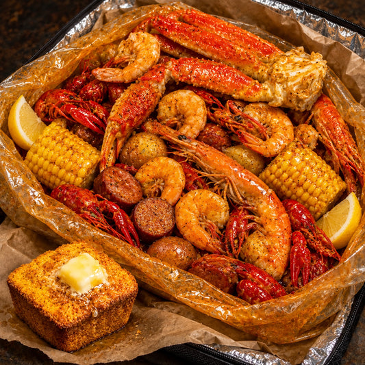 A close-up overhead shot of a seafood boil or crawfish boil served in a foil-lined tray, with shrimp, crawfish, corn, potatoes, sausage, lemon wedges, and a buttered cornbread on brown paper. The food is heavily seasoned with red and yellow spices.