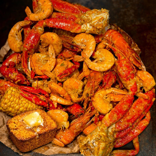 A close-up overhead shot of a seafood boil or crawfish and shrimp meal, featuring red-orange shrimp, crawfish, lobster legs, corn, and a piece of cornbread with butter on a dark pan or plate.