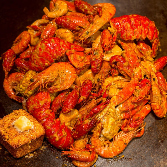 A close-up pile of cooked shrimp and crawfish in a spicy orange-red sauce, served on a dark plate with a small buttered bread cube at the lower left. Boil Baby Boil Seafood Boil Bags are always fresh and delivery friendly with insulated bag packaging.
