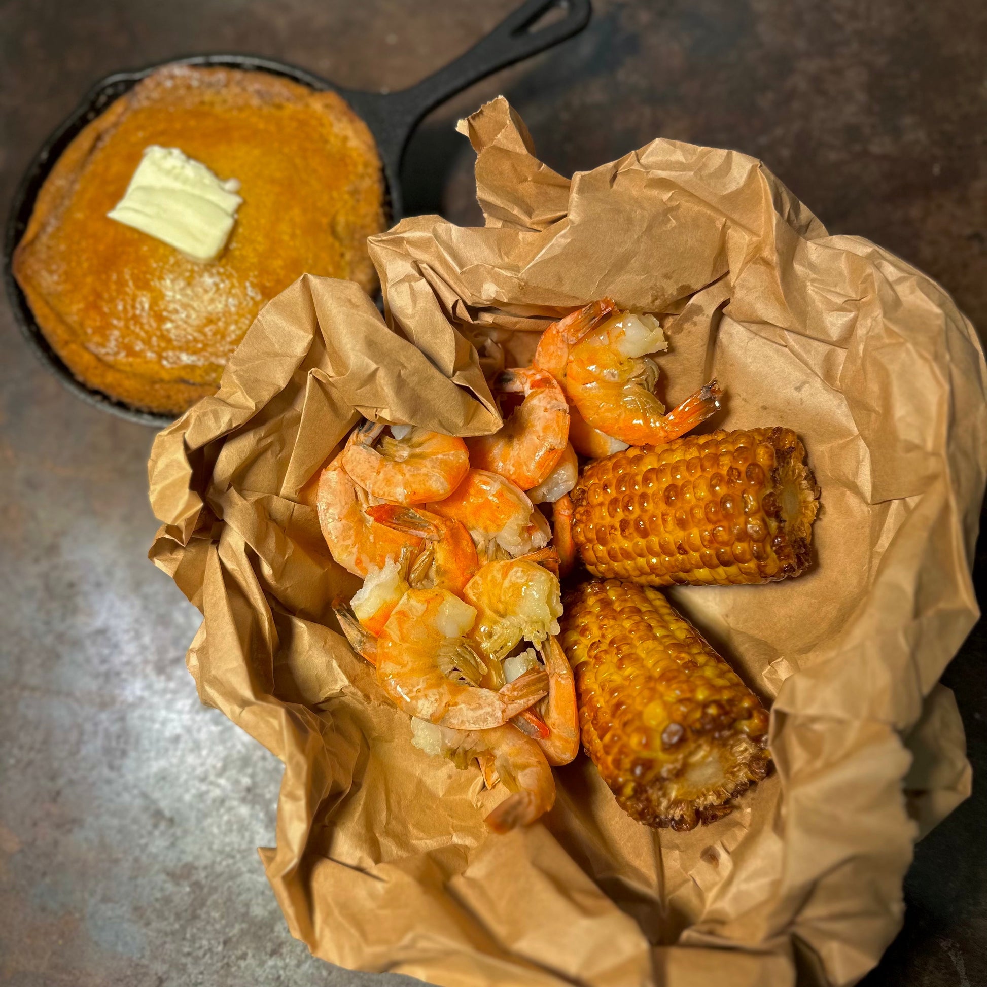 Juicy boiled shrimp with garlic, served alongside corn on the cob and a skillet of honey butter cornbread in a rustic Southern-style spread.
